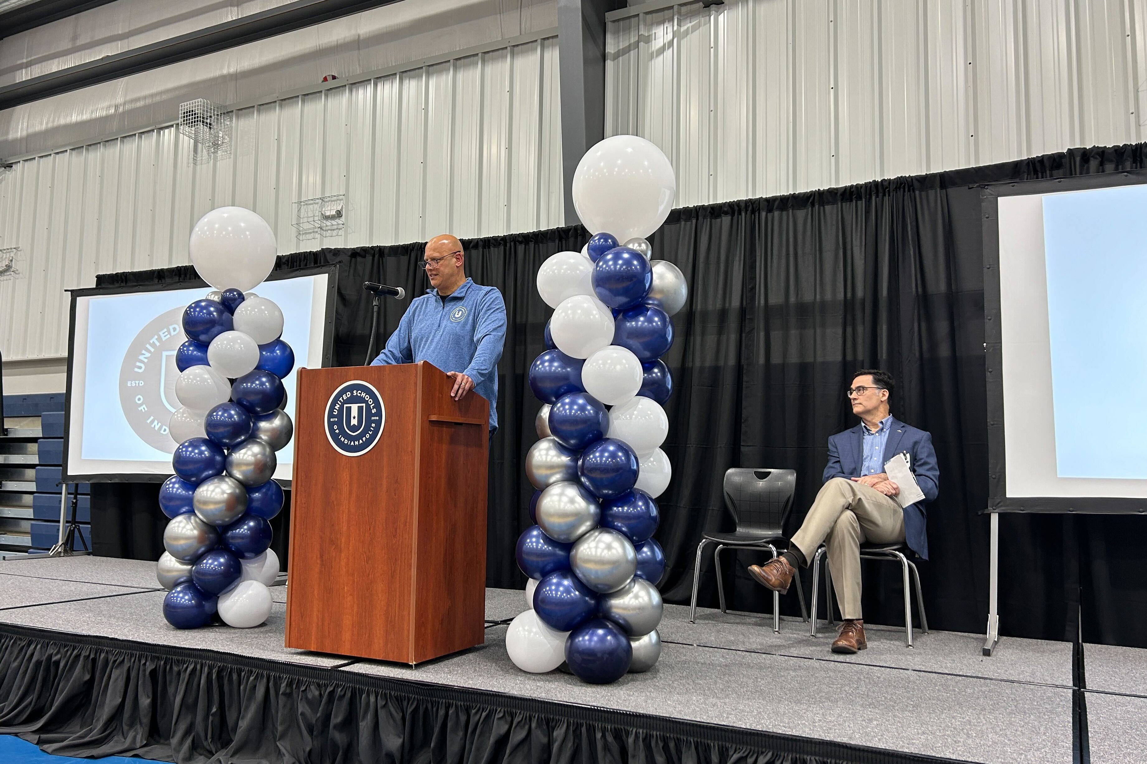 Two people on stage with white and blue balloons and a blue sheet for the background.