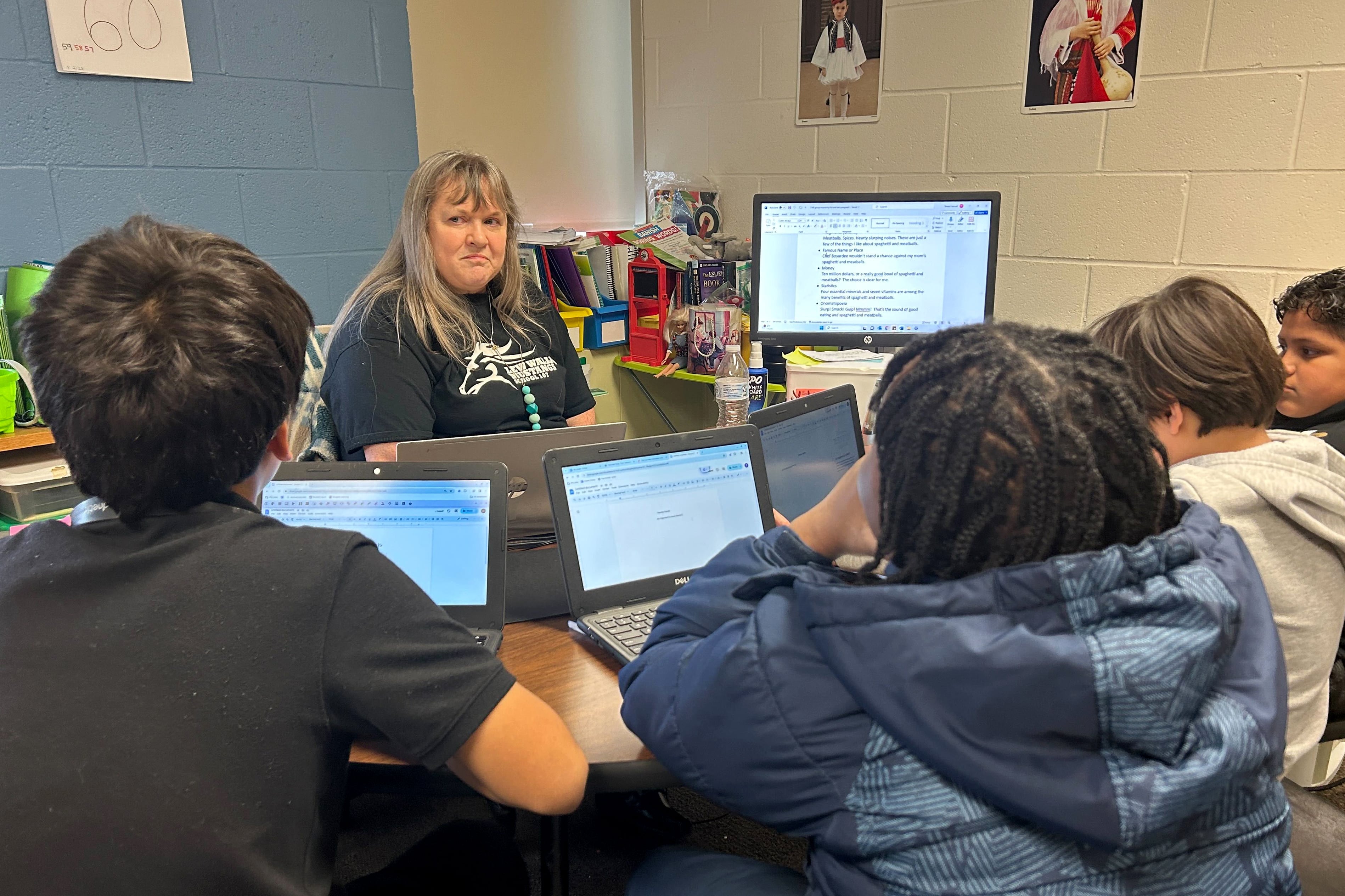 An adult with long white hair sits behind a desk in a classroom with four students sitting on the other side all working at computers.