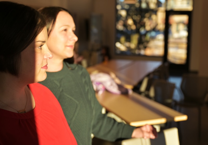 Two women in sweaters pose for a portrait in a beam of light in a building.