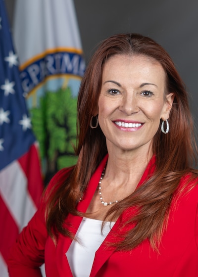 Image is an official portrait of Kirsten Baesler. She has long, brown hair and is wearing a red blazer and white blouse.