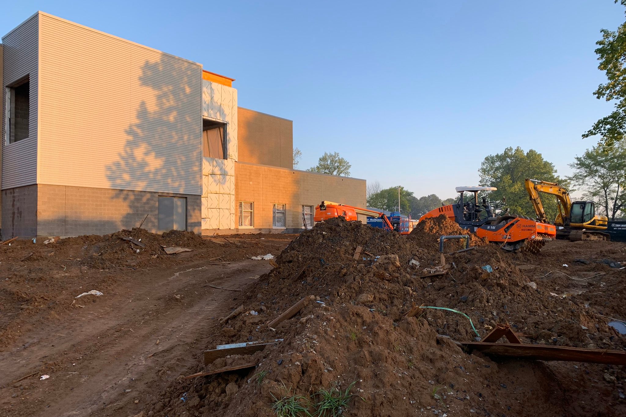 Orange and yellow bulldozers maneuver through a large pile of dirt under a blue sky outside of a new tan, two-story school building that is nearing completion.