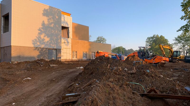 Orange and yellow bulldozers maneuver through a large pile of dirt under a blue sky outside of a new tan, two-story school building that is nearing completion.