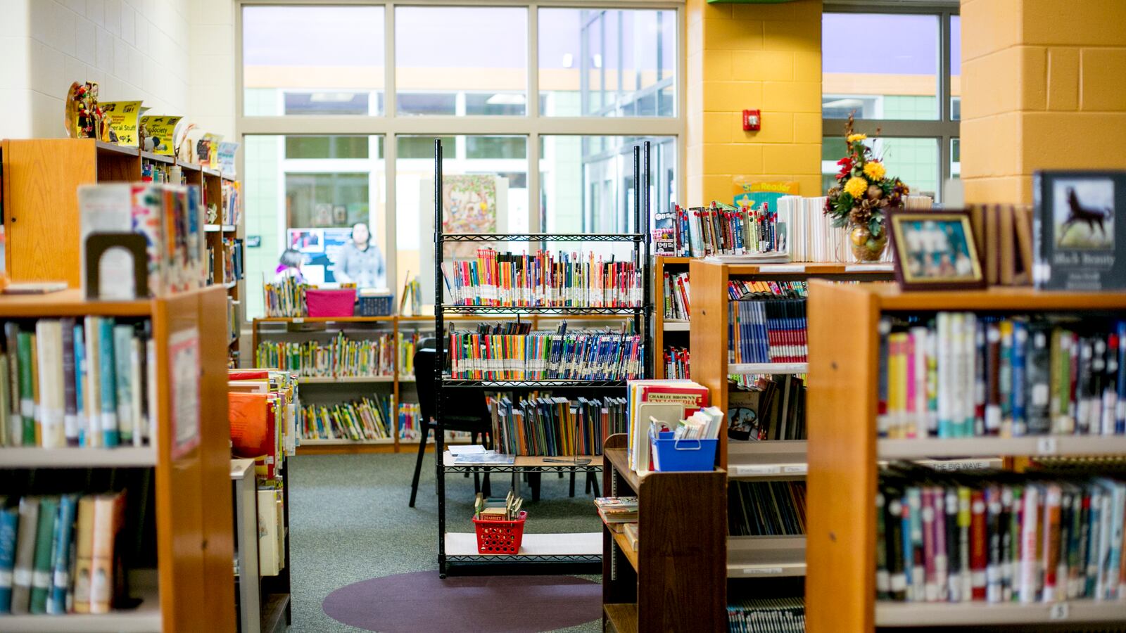 A library with shelves full of colorful books lining the walls with one black chair in the middle of the image. There is a large glass window looking out to another part of the school in the background.