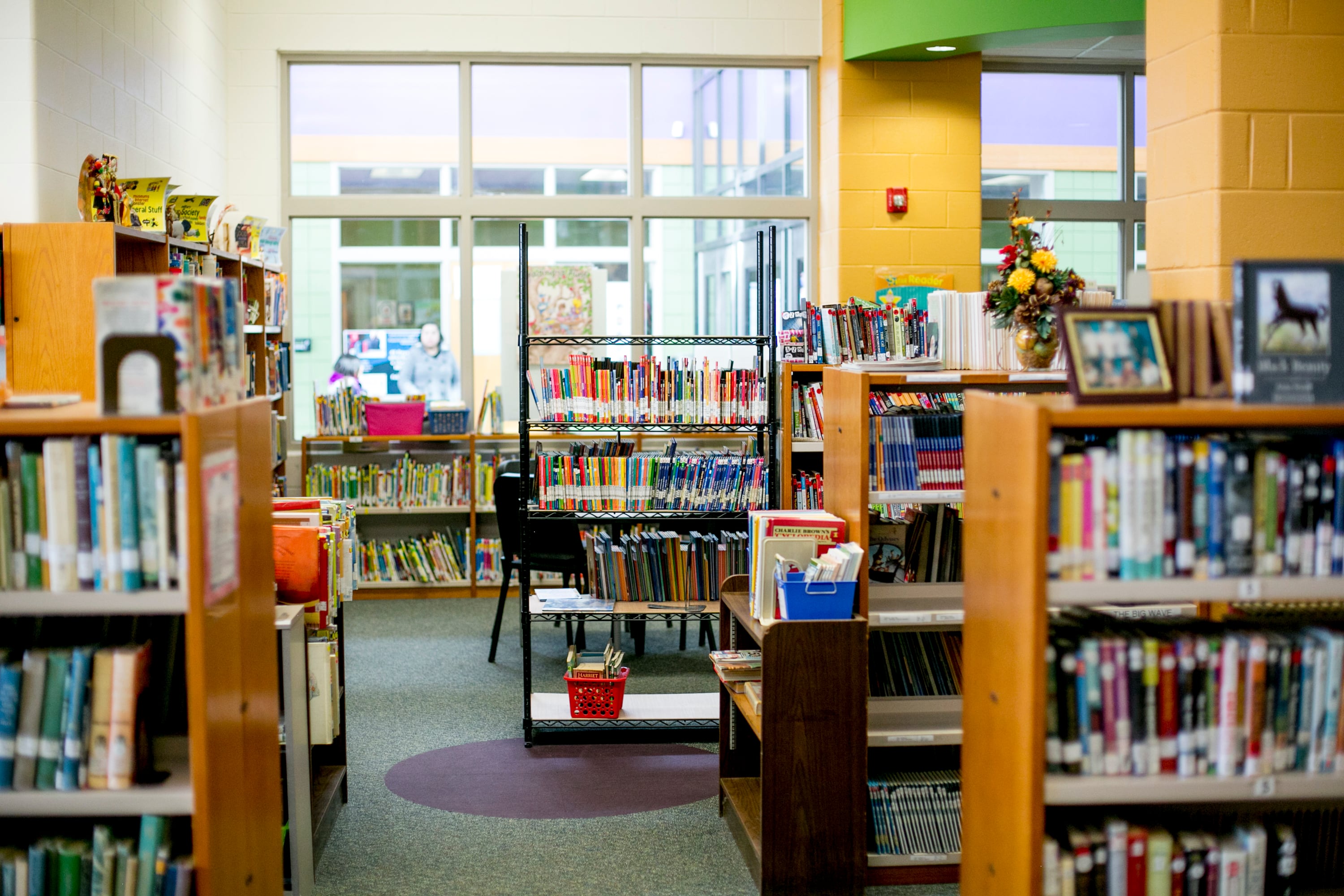 A library with shelves full of colorful books lining the walls with one black chair in the middle of the image. There is a large glass window looking out to another part of the school in the background.