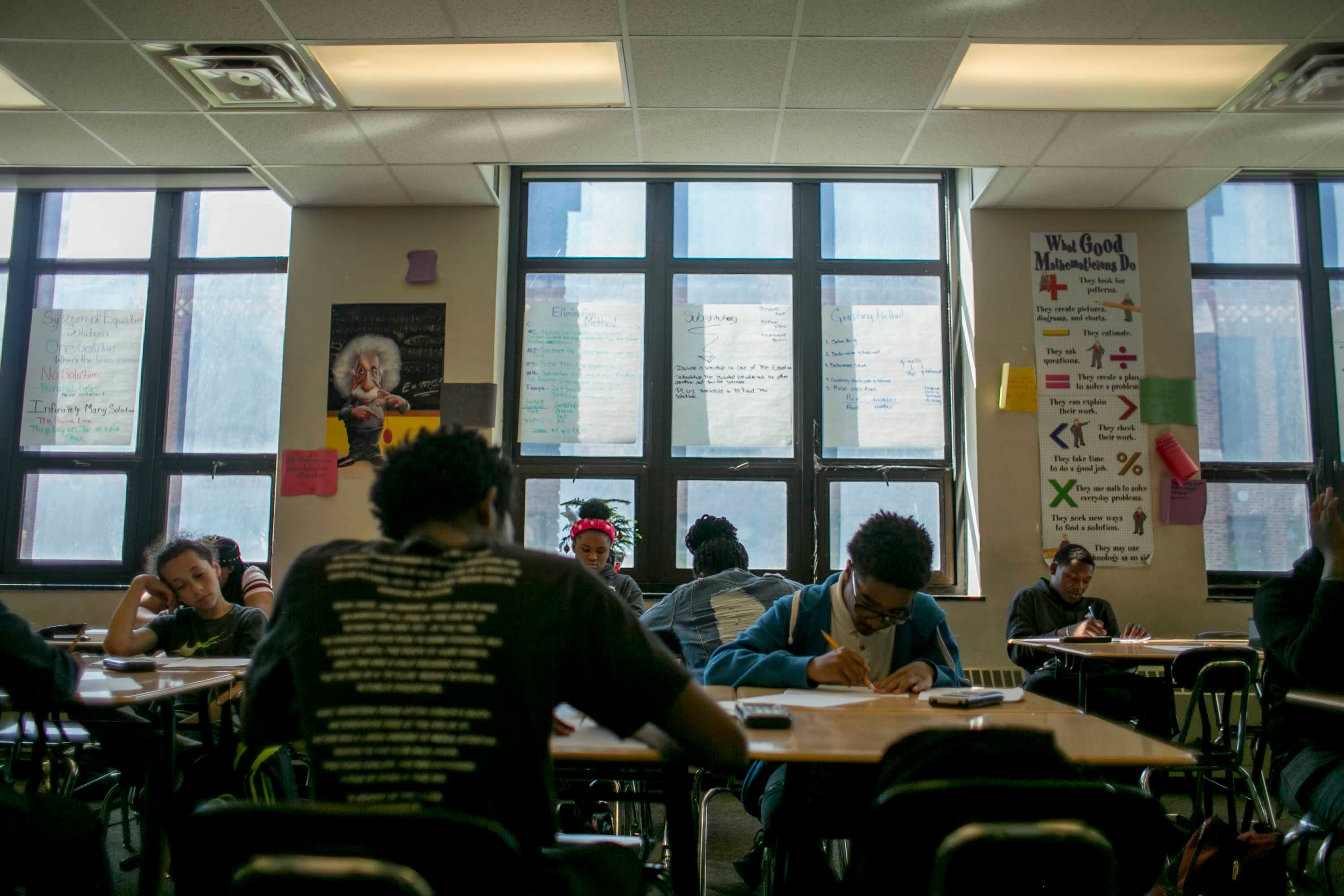 Several students sit at desks in a classroom with large windows.