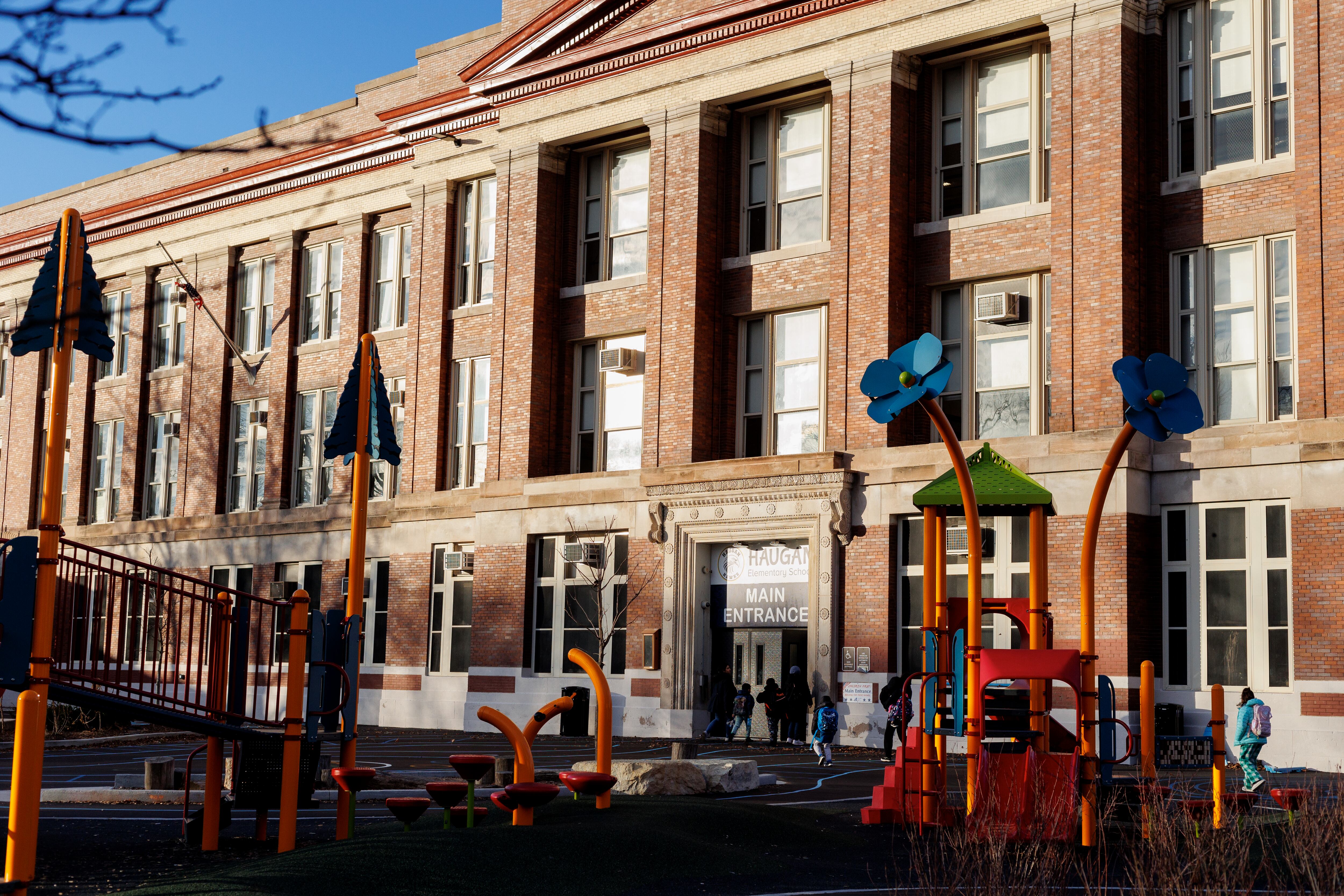 The front entrance of a large school building with students walking toward the door. There is a playground in the foreground.