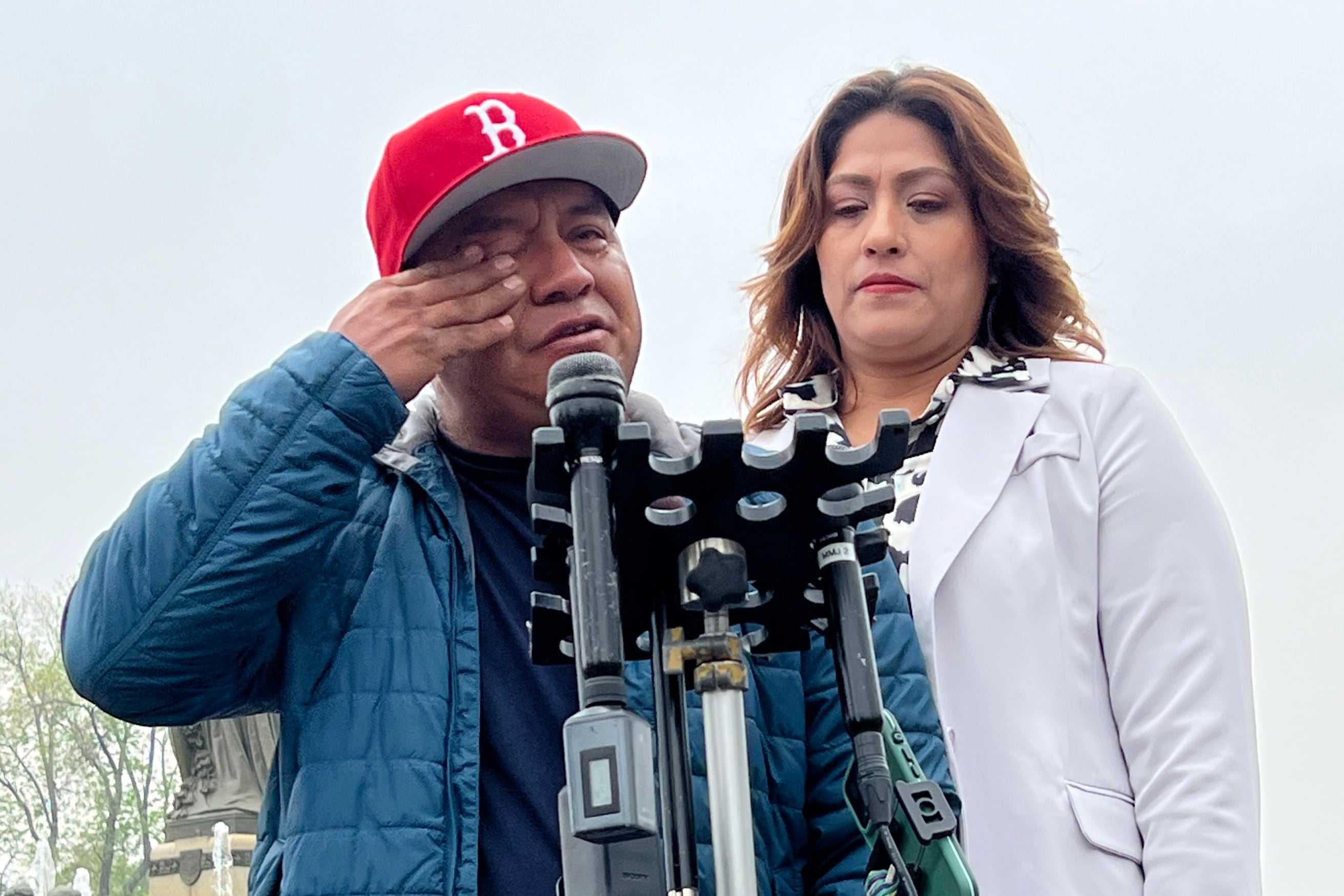 a man in a blue jacket and red cap wipes his eye while talking at a microphone next to a woman in a white suit.