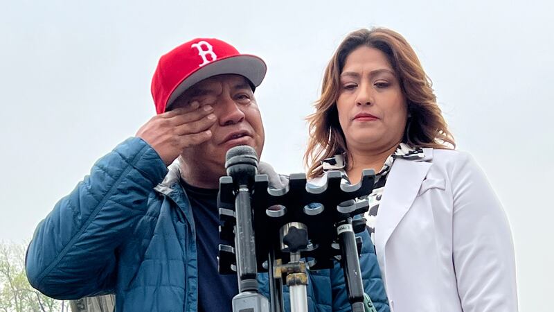 a man in a blue jacket and red cap wipes his eye while talking at a microphone next to a woman in a white suit.