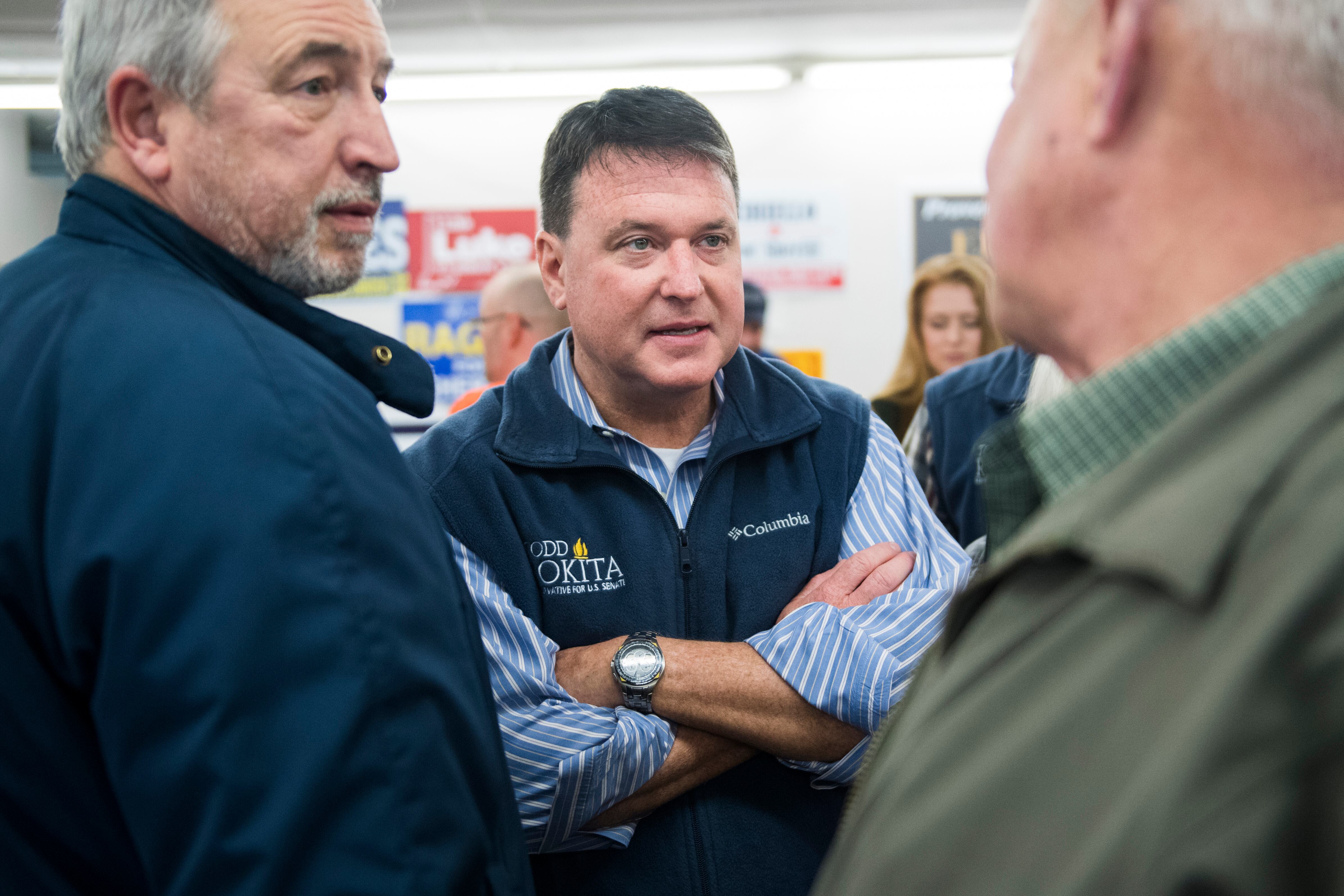 A man in a blue vest and blue shirt stands with arms folded between two other men.