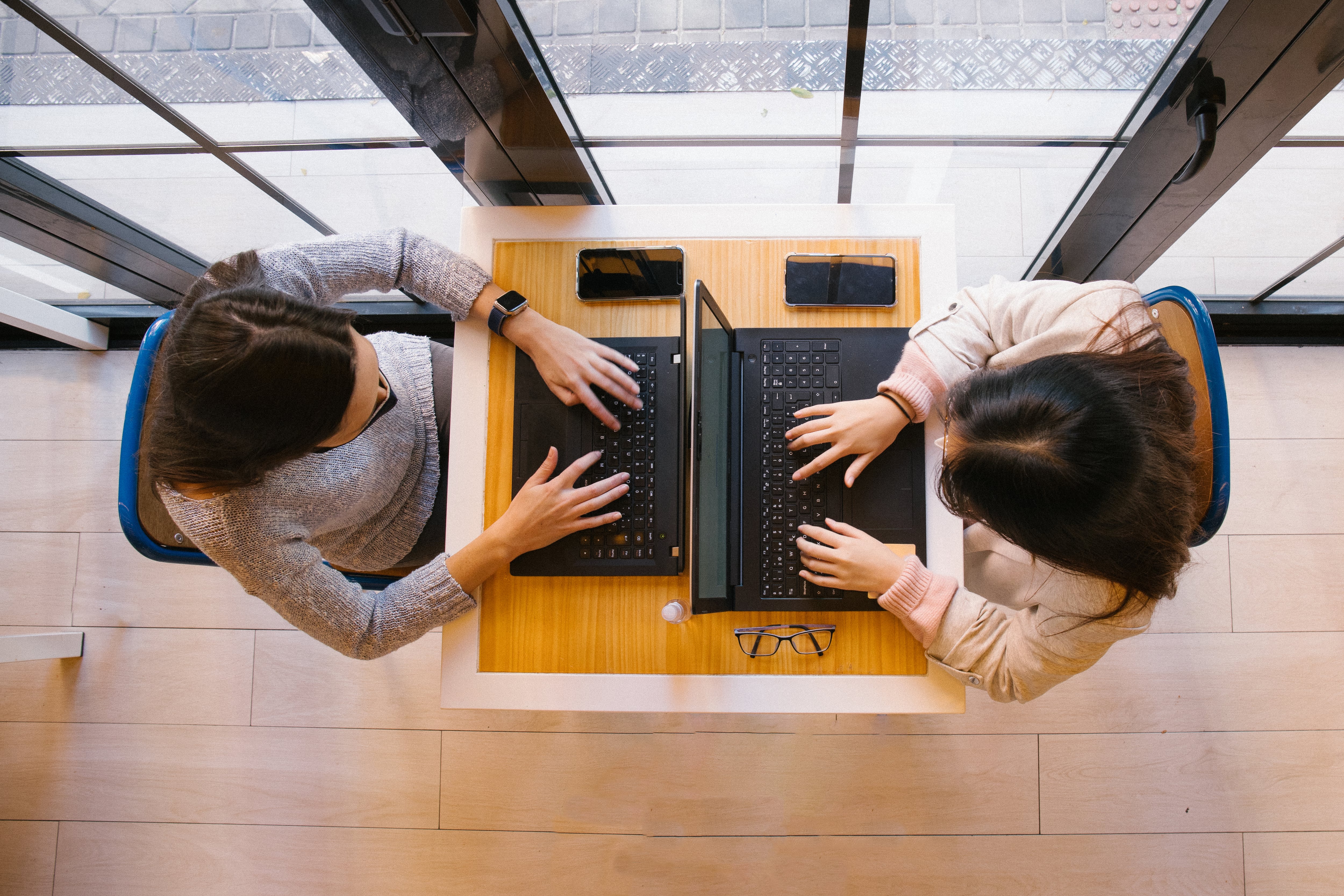 Two young women work at their laptops in front of a window.