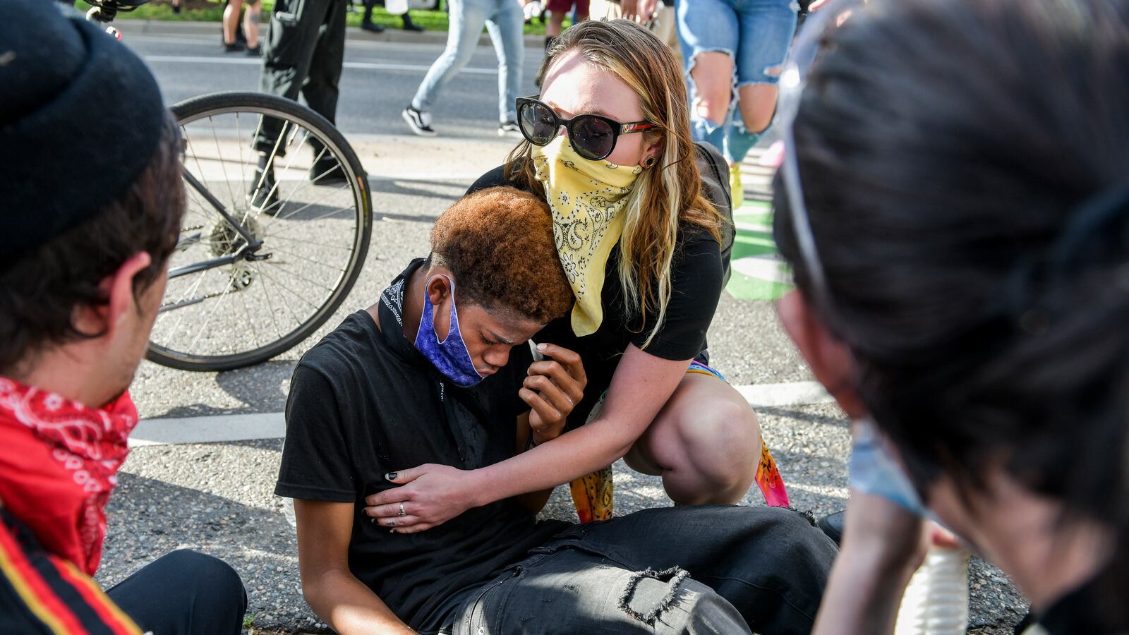 A woman wearing a yellow bandana and black shirt consoles a young man wearing a black shirt and jeans who is under duress. There are two people in the foreground, one with a red bandana.