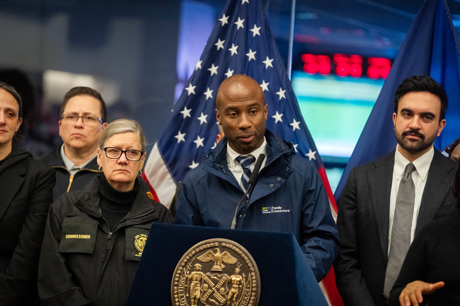 A photograph of a Black man standing behind a podium while speaking during a press conference while a group of people stand next to him.