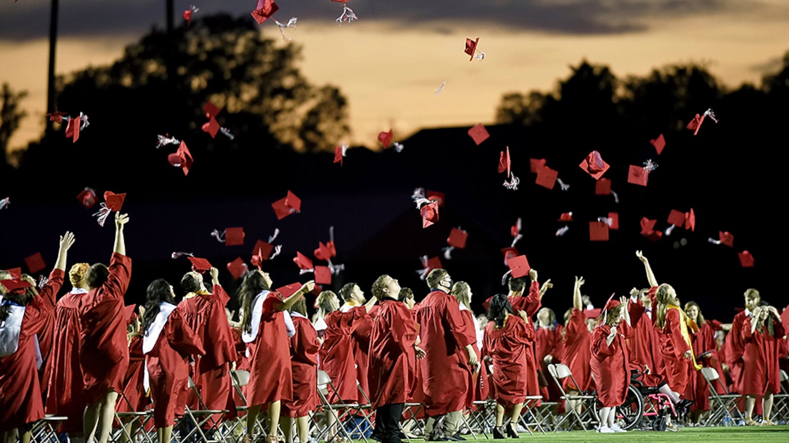 Graduation seniors in red gowns toss their graduation mortarboards into the air against a dark evening sky.