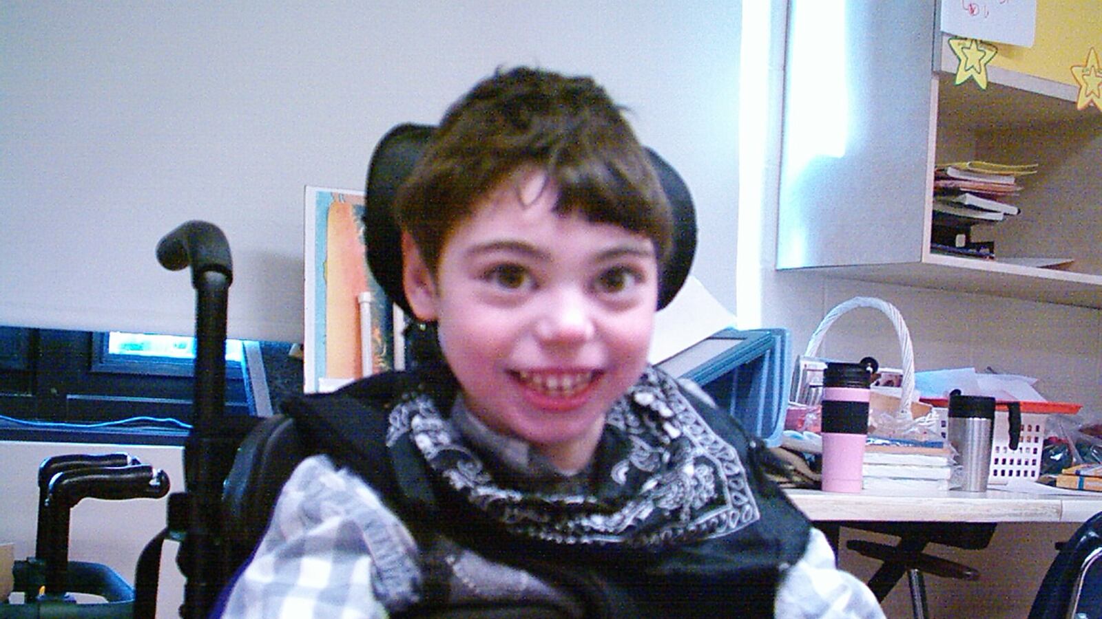 A little boy with brown hair smiles. He sits in a Black wheelchair. He is wearing a gray and white shirt.