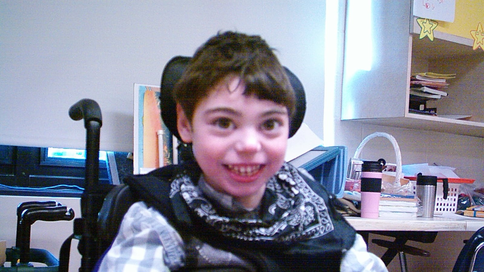 A little boy with brown hair smiles. He sits in a Black wheelchair. He is wearing a gray and white shirt.