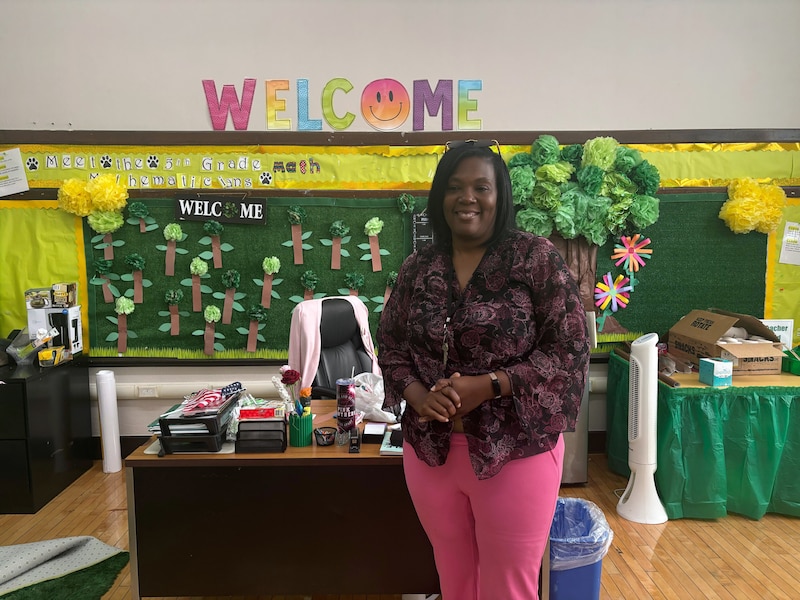 A photograph of a Black woman wearing a dark blouse and pink pants poses for a photograph at the front of a colorful classroom.
