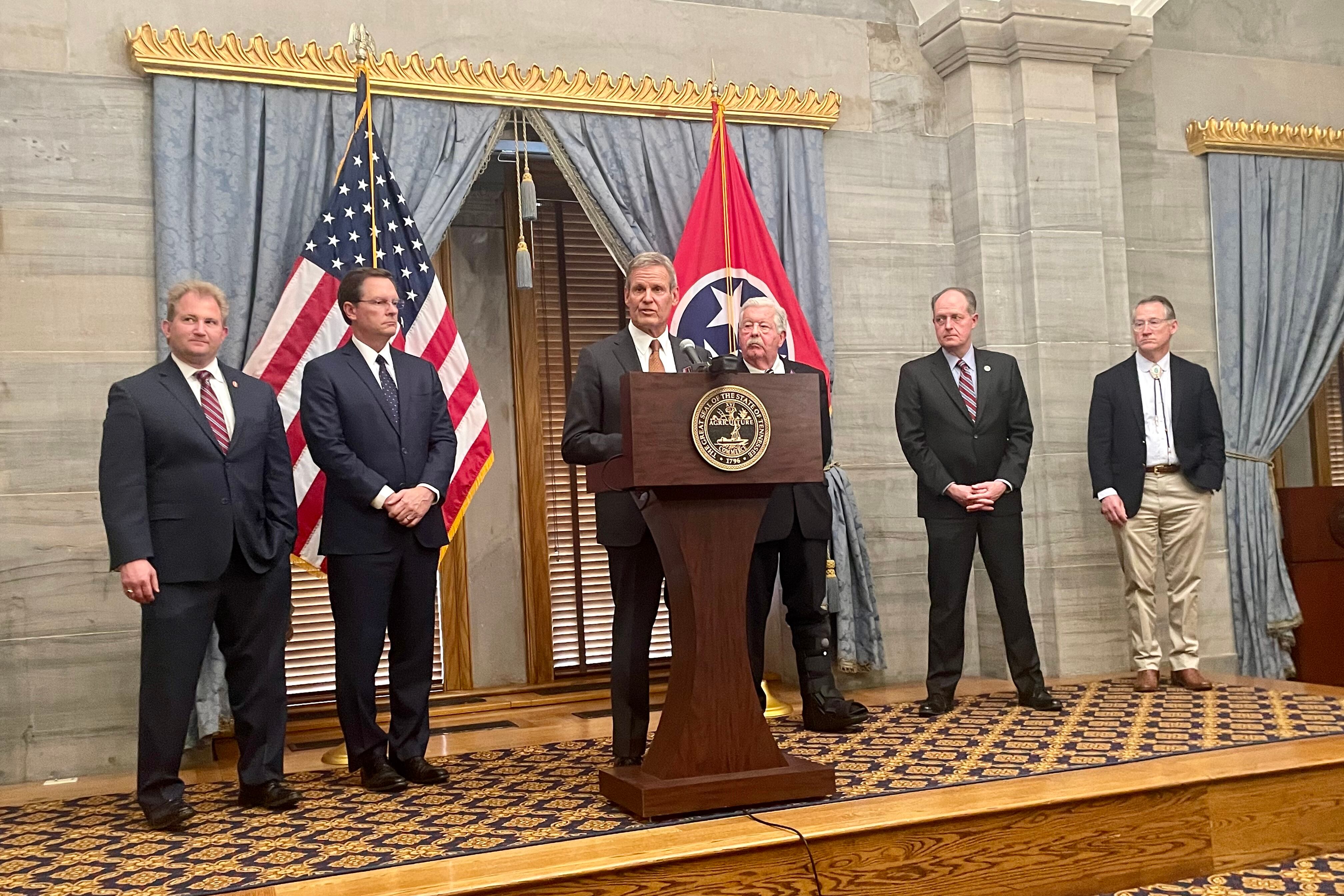 Six men wearing suits stand side by side while the person in the middle stands behind a wooden podium there is an American flag and a Tennessee state flag in the background.