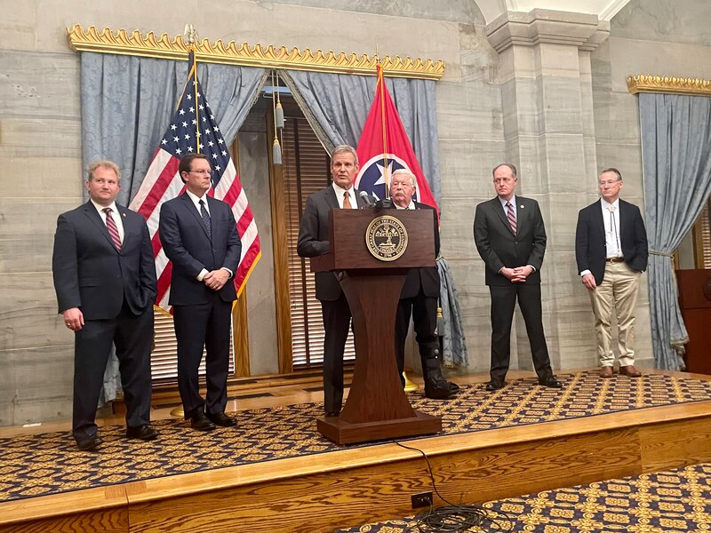 Six men wearing suits stand side by side while the person in the middle stands behind a wooden podium there is an American flag and a Tennessee state flag in the background.