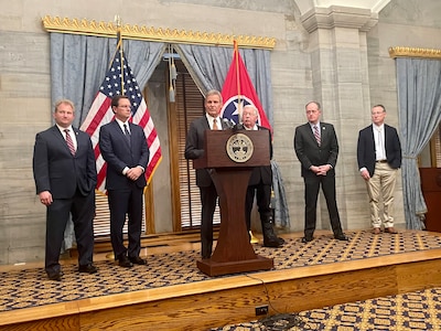 Six men wearing suits stand side by side while the person in the middle stands behind a wooden podium there is an American flag and a Tennessee state flag in the background.