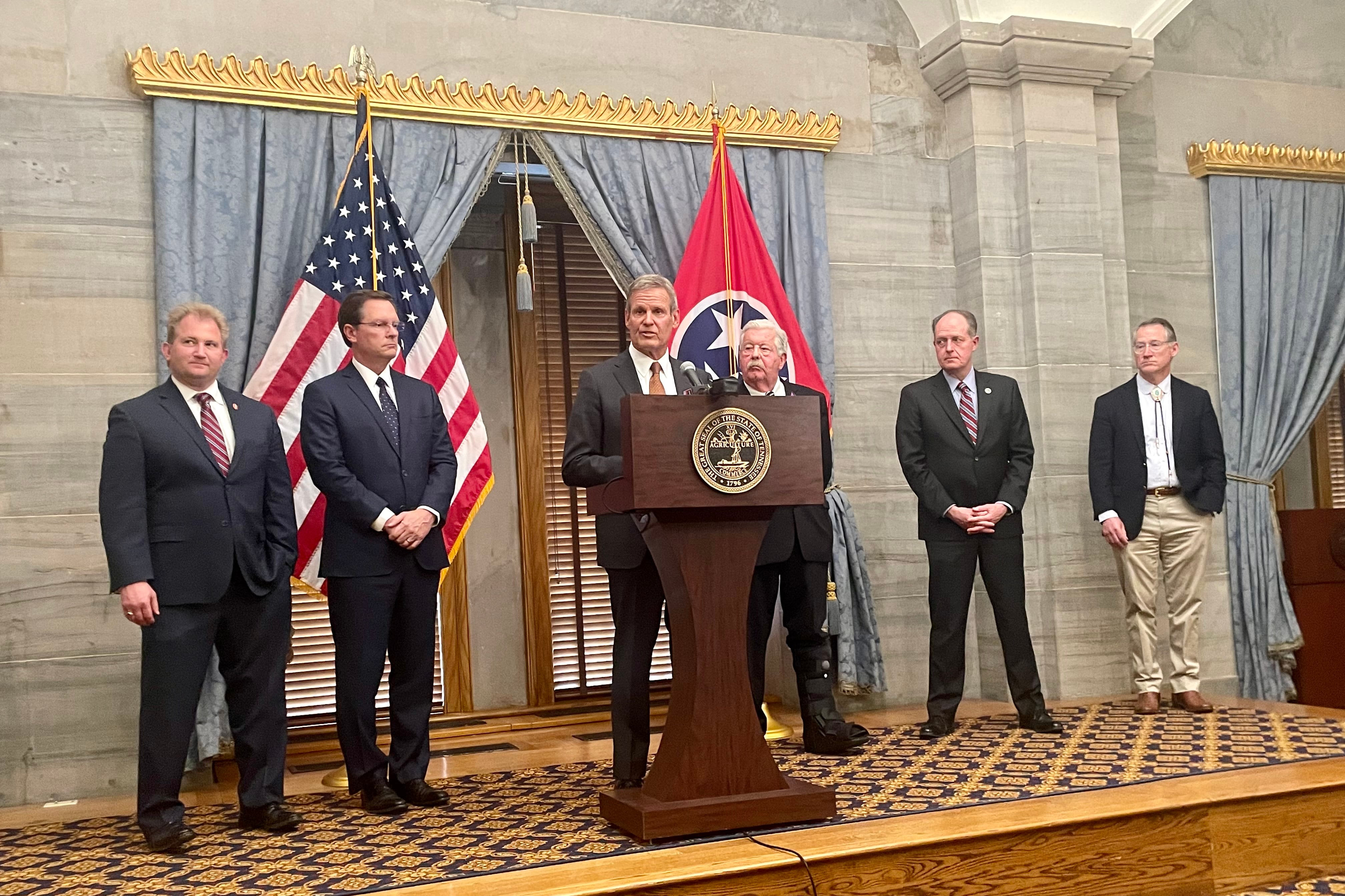 Six men wearing suits stand side by side while the person in the middle stands behind a wooden podium there is an American flag and a Tennessee state flag in the background.