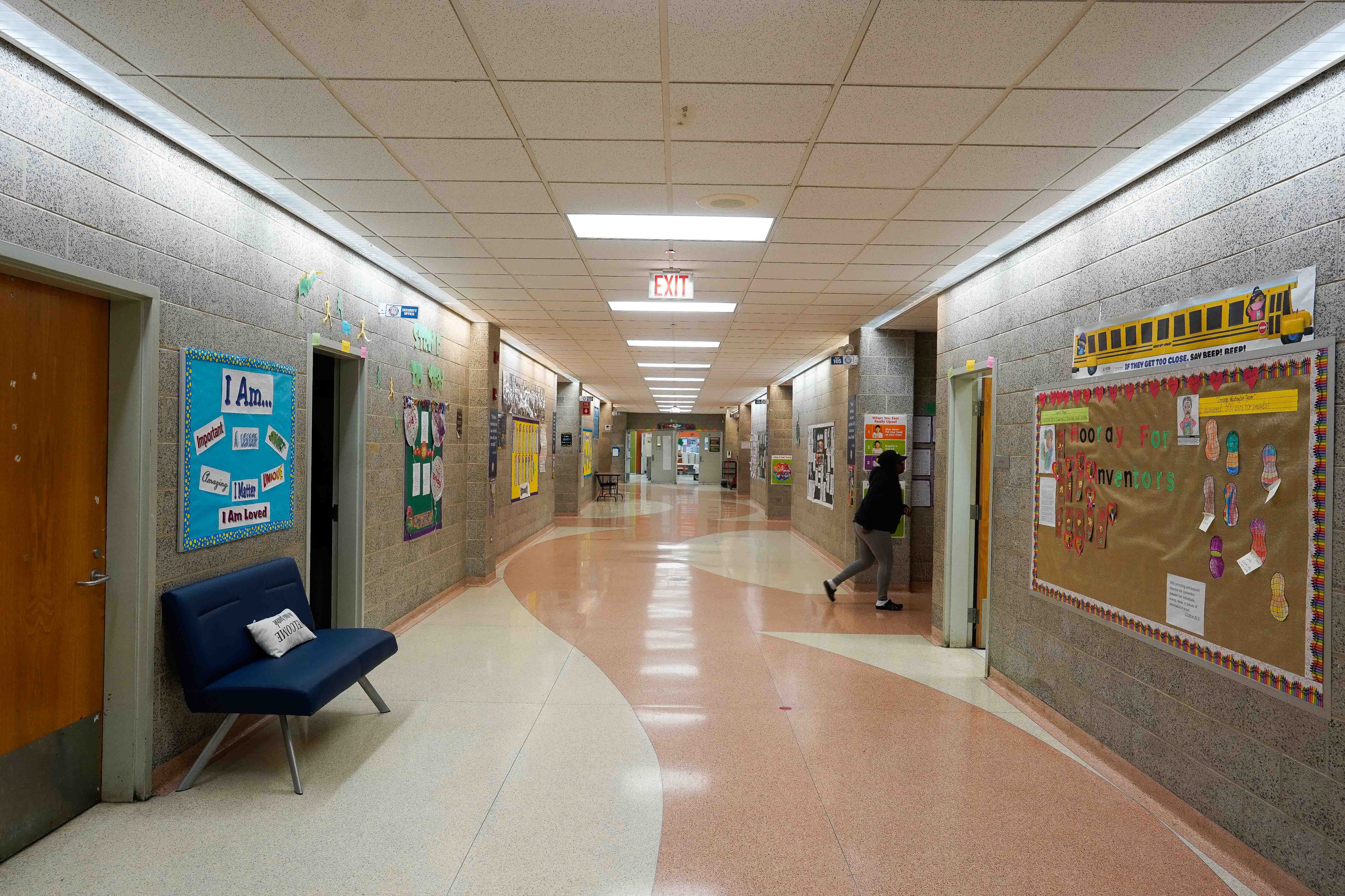 A school hallway with one student walking into a classroom door. The walls are covered in bulletin boards and art.