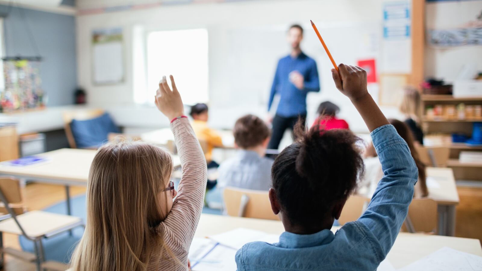 two students raise their hands in the foreground while sitting at a desk while in the background a teacher stands in front of a whiteboard