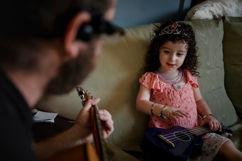 A photograph of a young girl with dark brown hair and a pink shirt sitting on a green couch with her dad in the foreground.