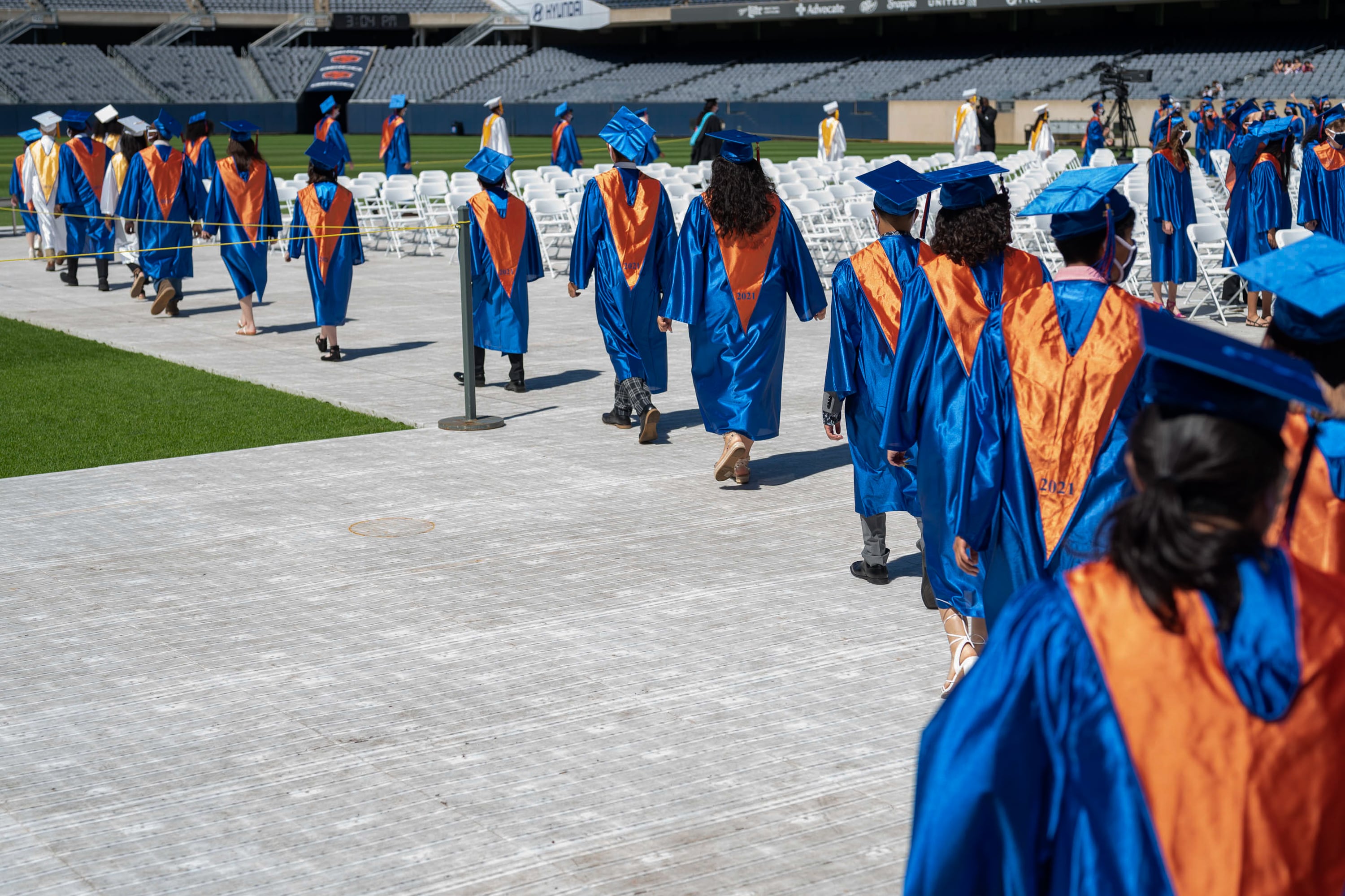 A line of high school graduates wearing orange and blue gowns walk down a path to their seats ahead of a ceremony.