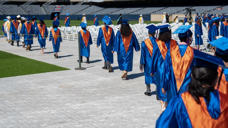 A line of high school graduates wearing orange and blue gowns walk down a path to their seats ahead of a ceremony.