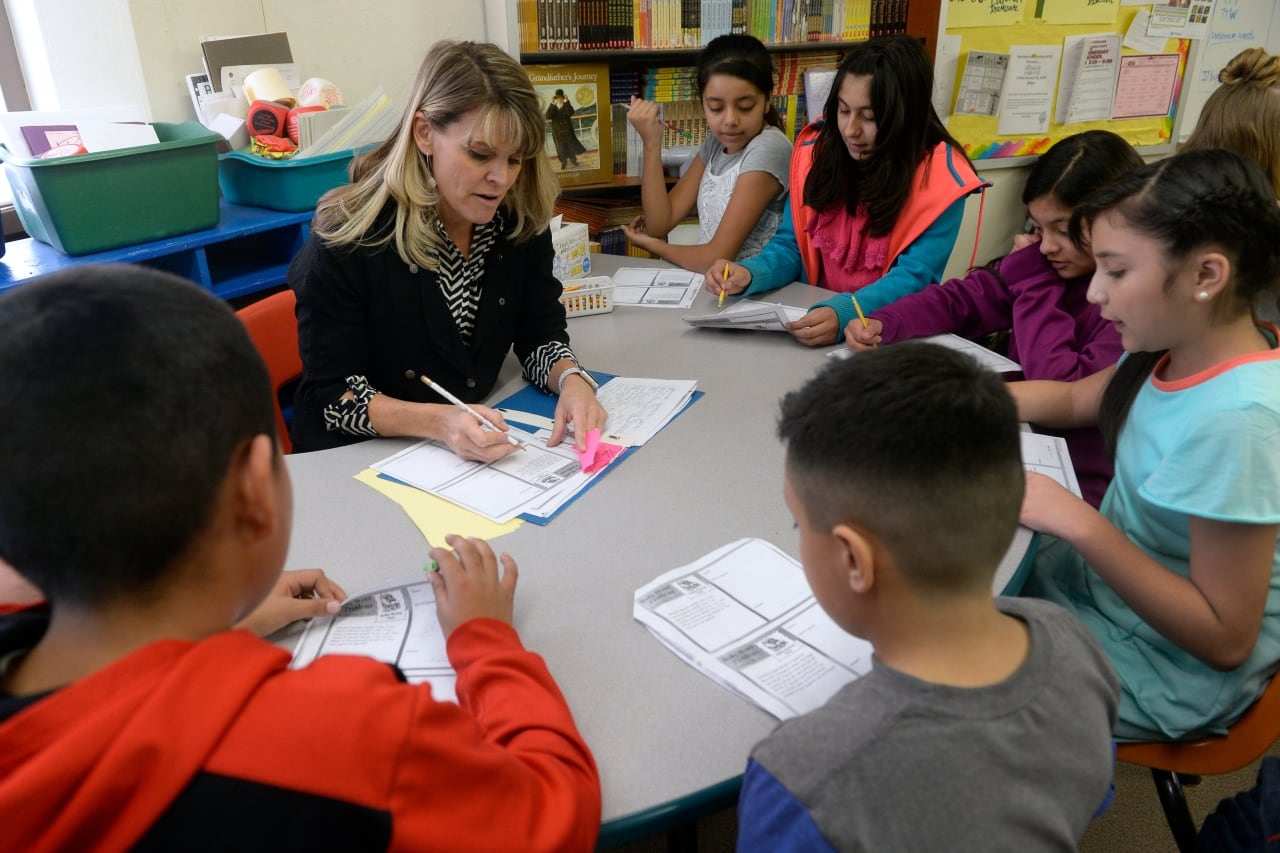 A teacher sits at a horseshoe desk working with six elementary-age children. They have papers on the desk, and the students are following along.