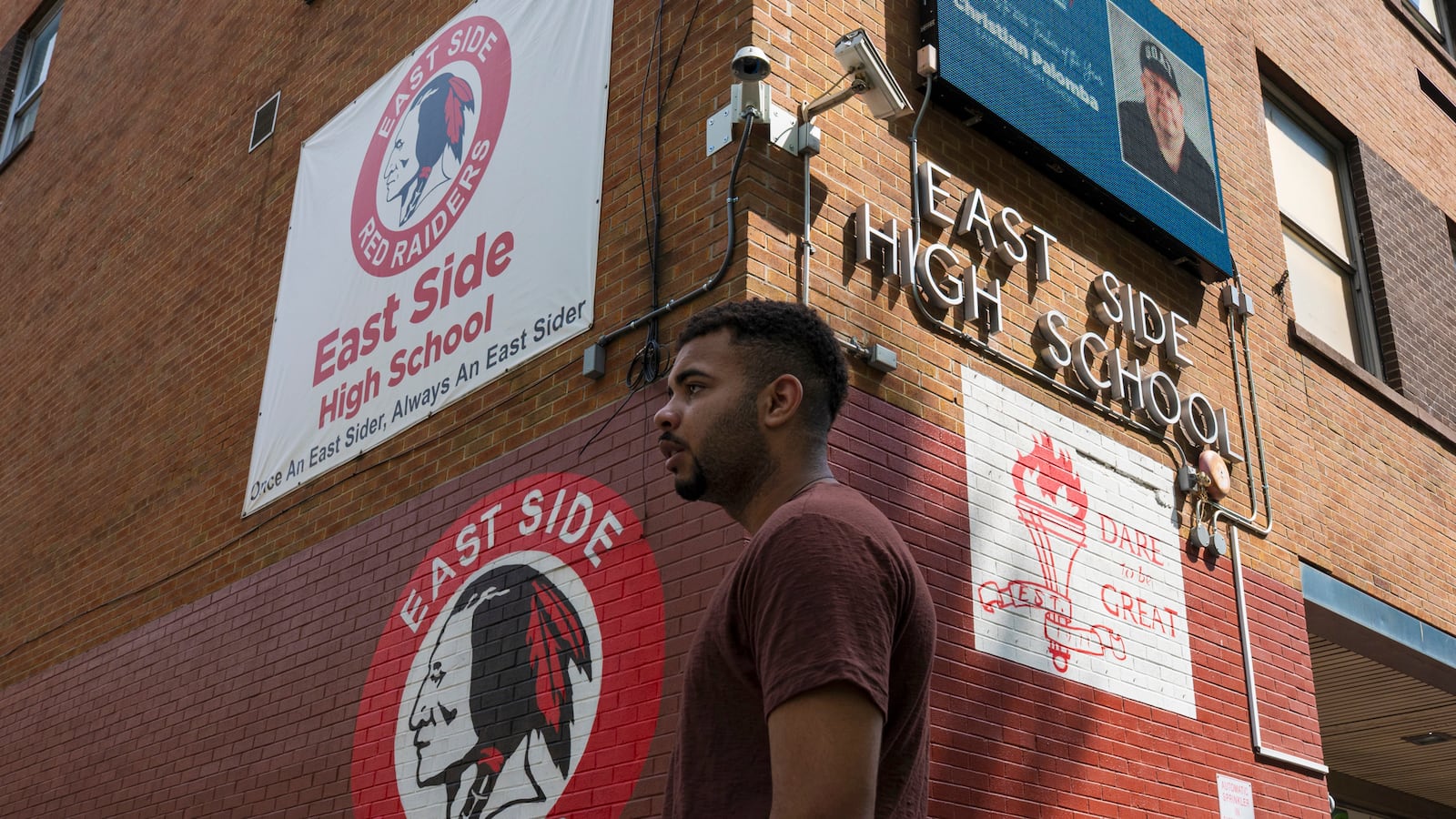 A high school student in a red shirt stands outside of his high school brick building.