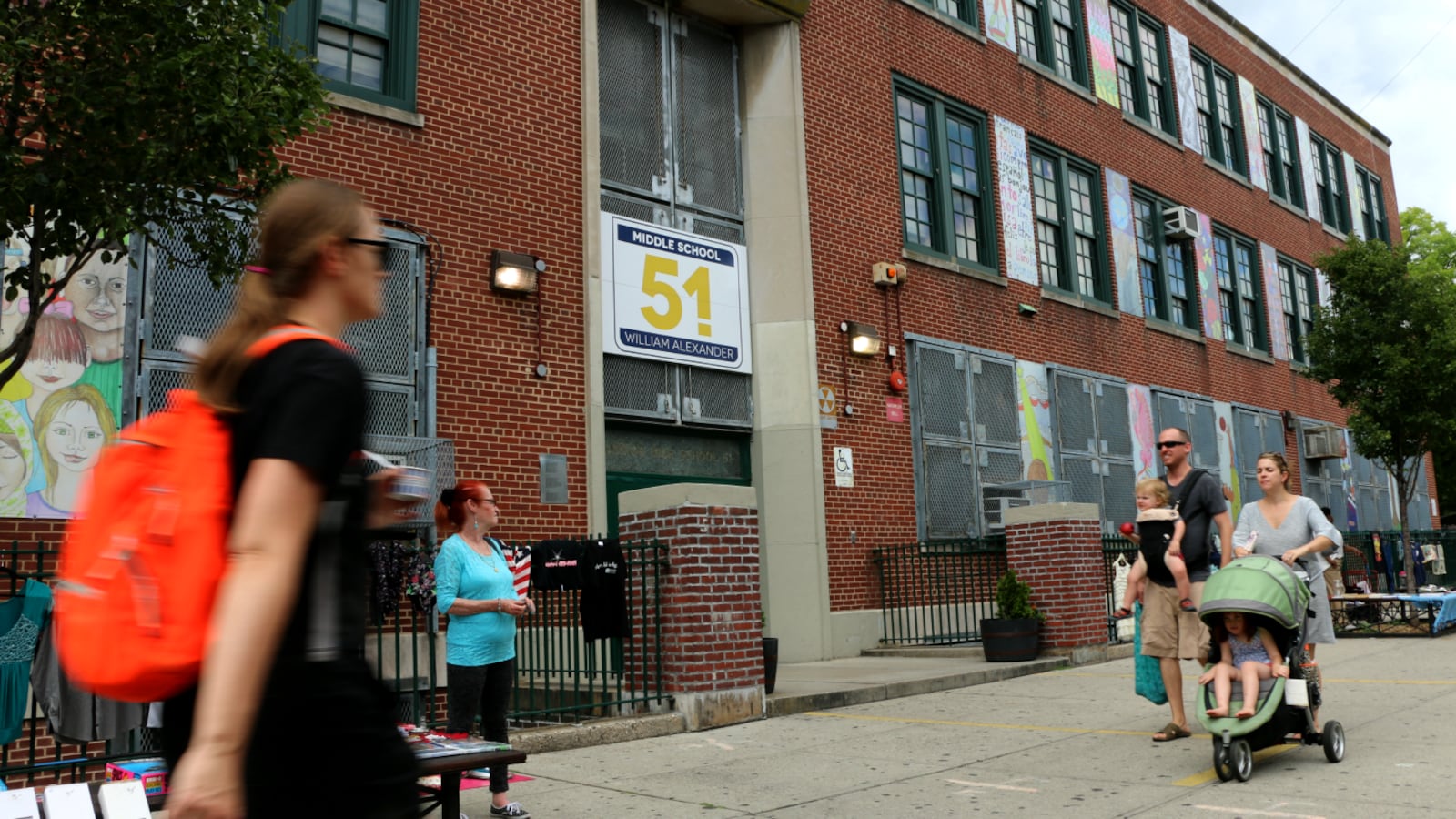 A person wearing an orange backpack walks past a brick school with a few other people in the background.