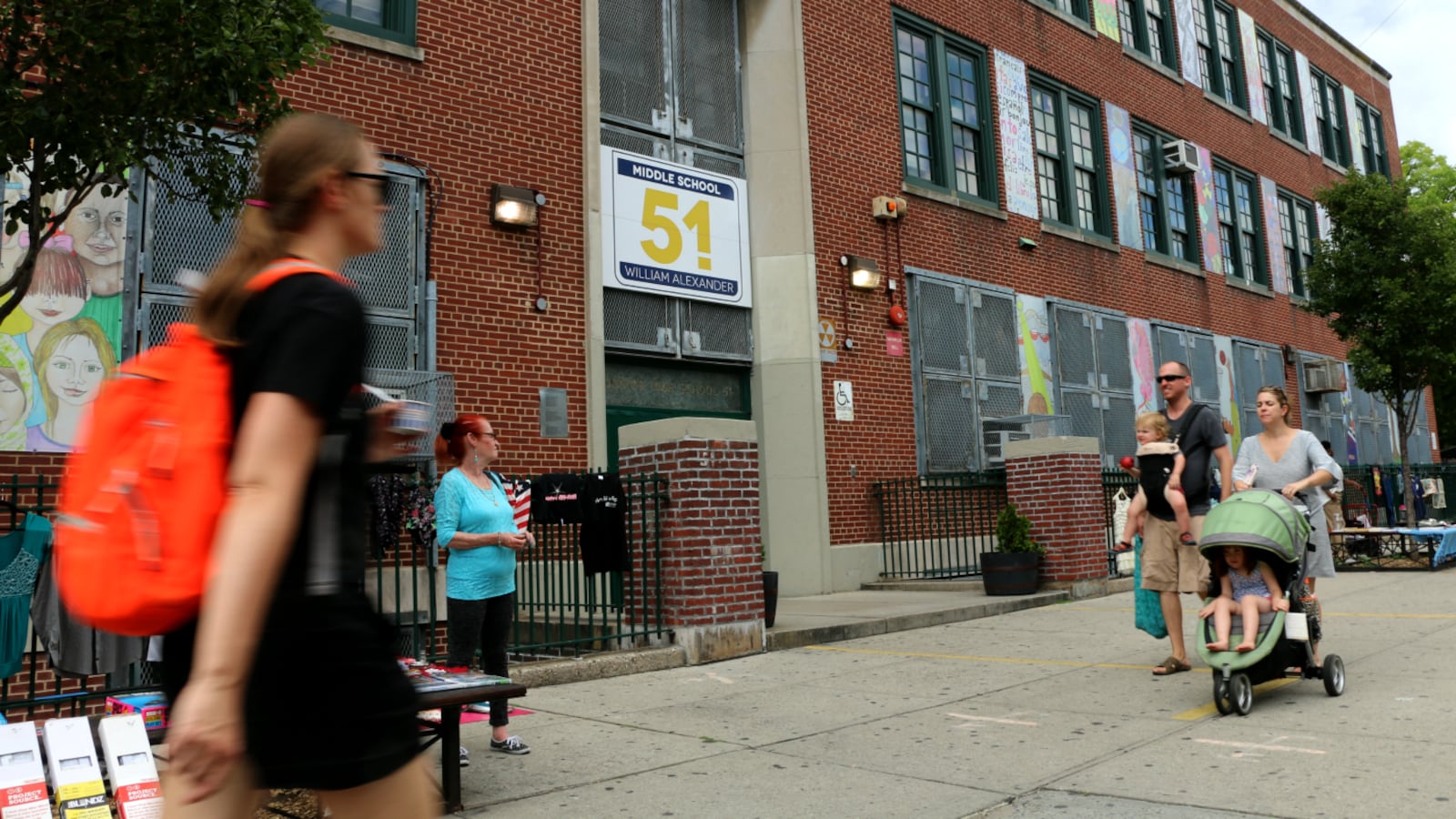 A person wearing an orange backpack walks past a brick school with a few other people in the background.