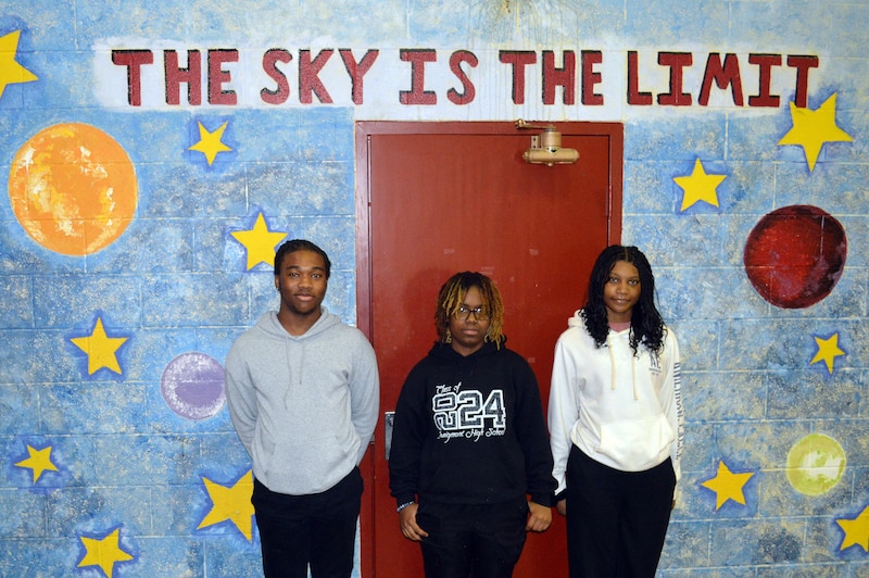 Three high school students stand side by side to pose for a portrait there is a mural in the background that reads "The Sky is the Limit" in red words.