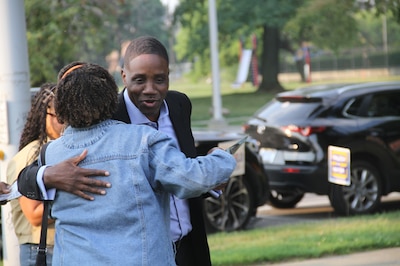 A photograph of a Black man in a suit hugs a woman outside with trees and cars in the background.