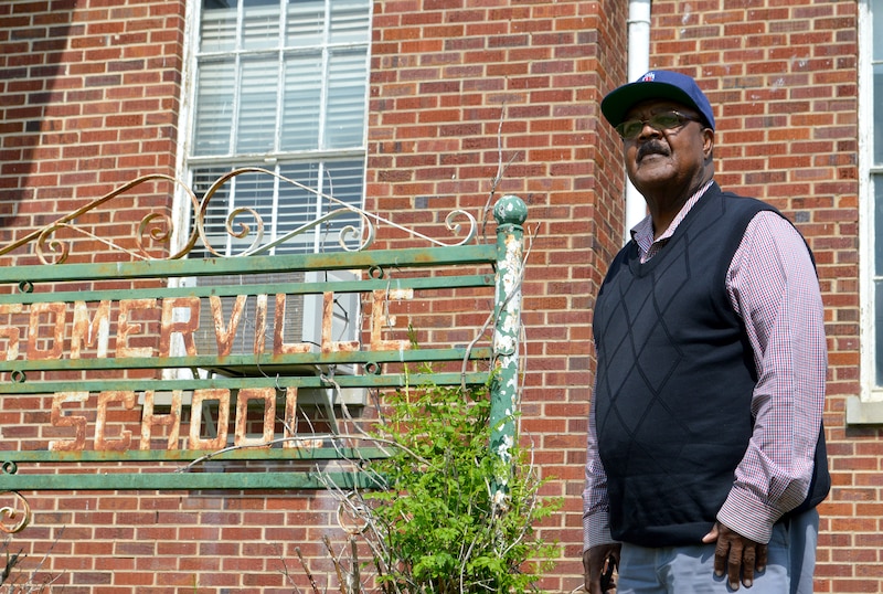 A man wearing a ball cap and a red and white striped shirt with a great sweater vest poses for a portrait in front of a red brick building.