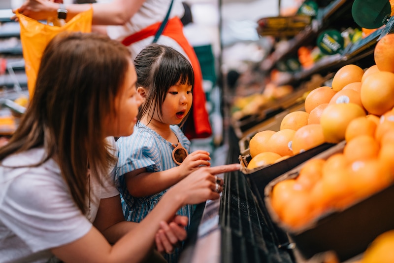 A mom and her daughter look at a bin full of large oranges in a grocery store with a person walking by in the background.