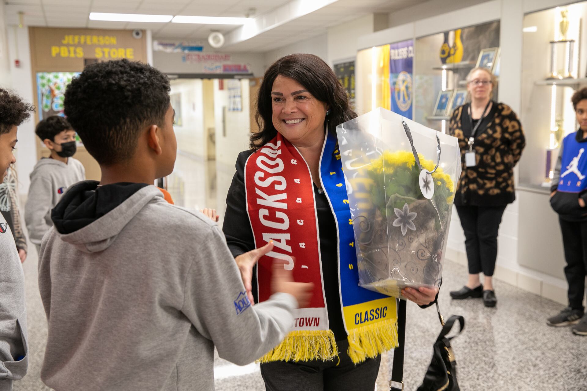 Carmen Ayala wears red and blue slash while carrying yellow flowers in one hand and shaking a students hand with the other. There are students and teachers in the hallways to welcome her and she’s at a school in Elgin’s U-46.