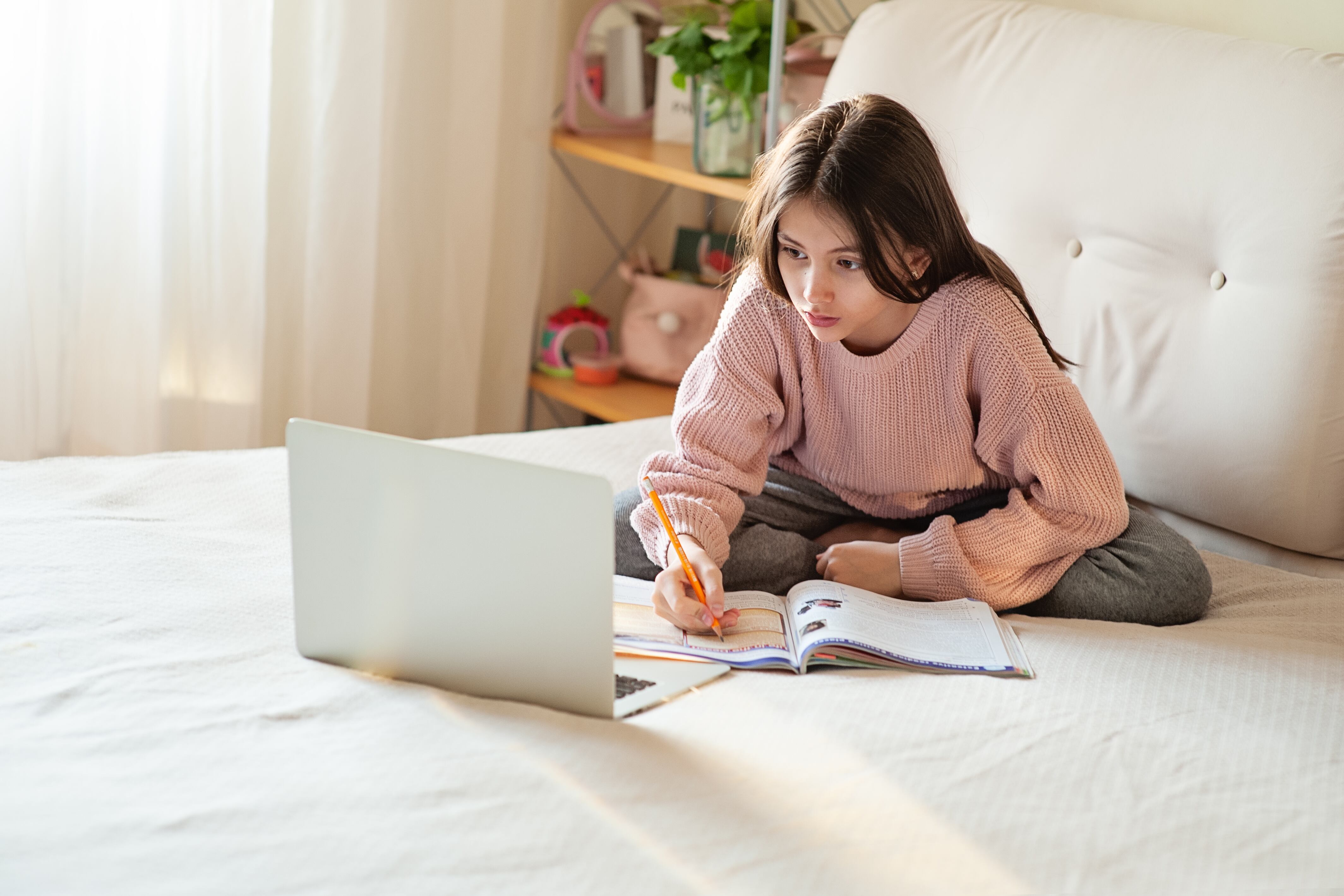 A young girl studies on a laptop and worksheet while sitting on her bed.
