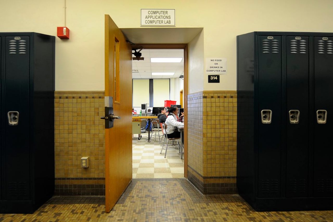 Two students sit in a classroom in the background with lockers and a classroom door in the foreground.