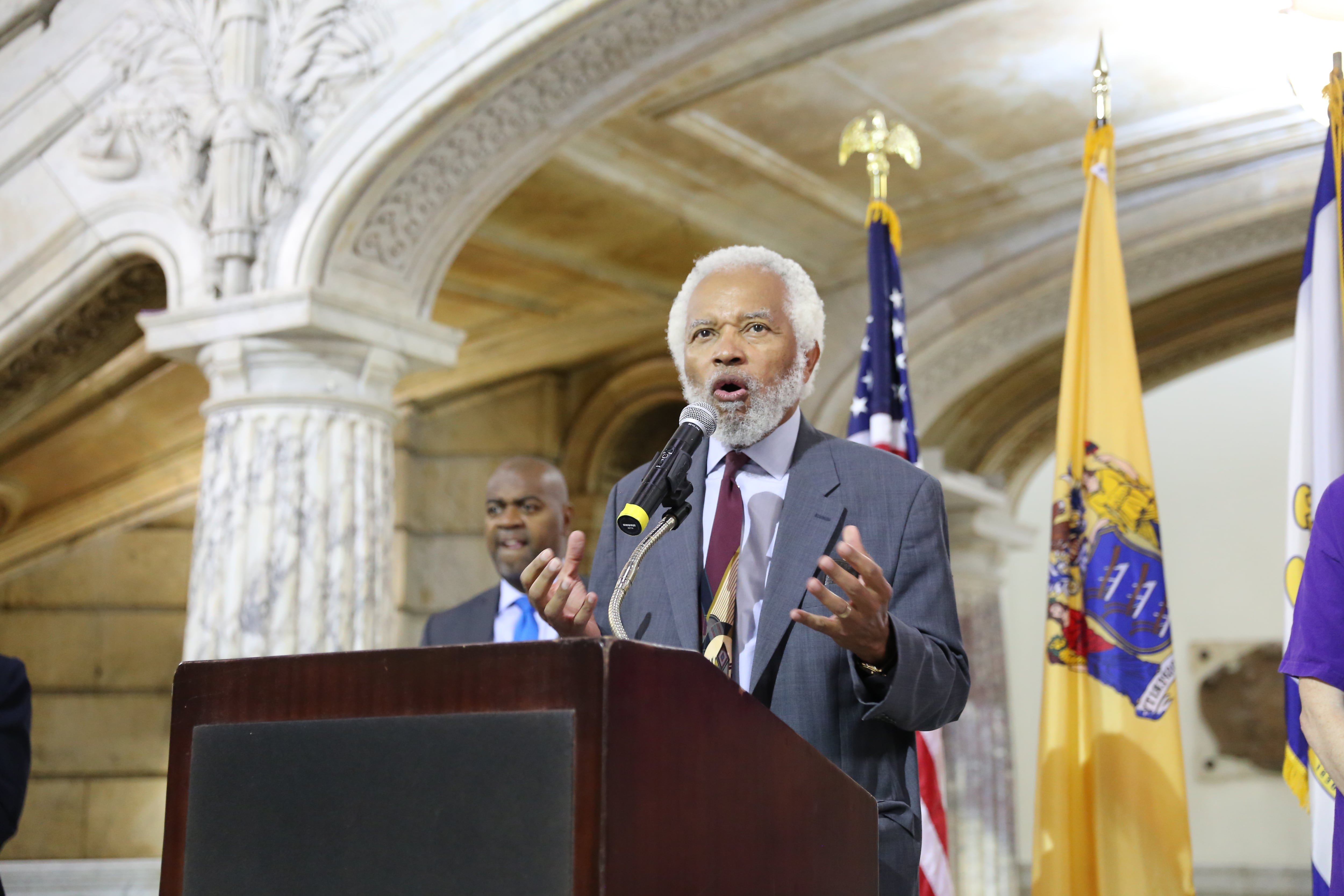 Junius Williams, in a gray suit and tie, gestures with his hands while standing at a lectern with flags behind him in a portico at Newark City Hall.