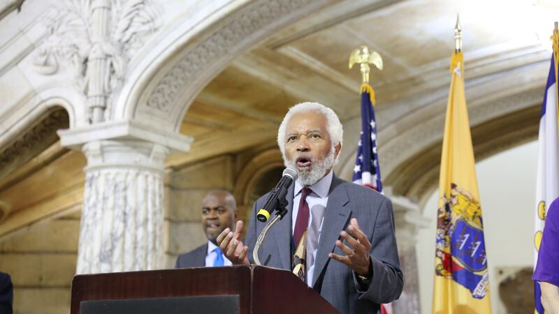 Junius Williams, in a gray suit and tie, gestures with his hands while standing at a lectern with flags behind him in a portico at Newark City Hall.