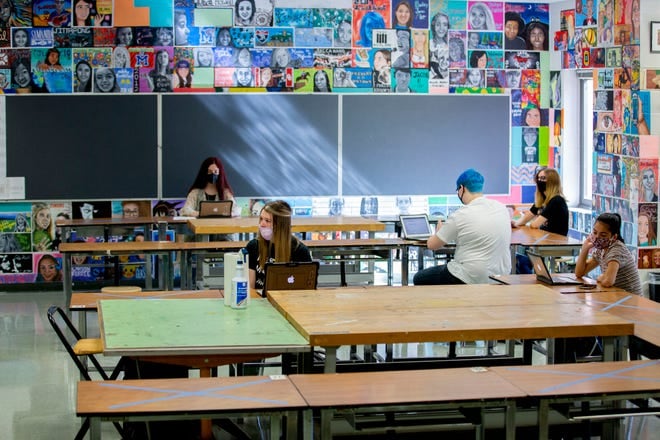 A teacher and students sit spread out in a room with laptops and masks.