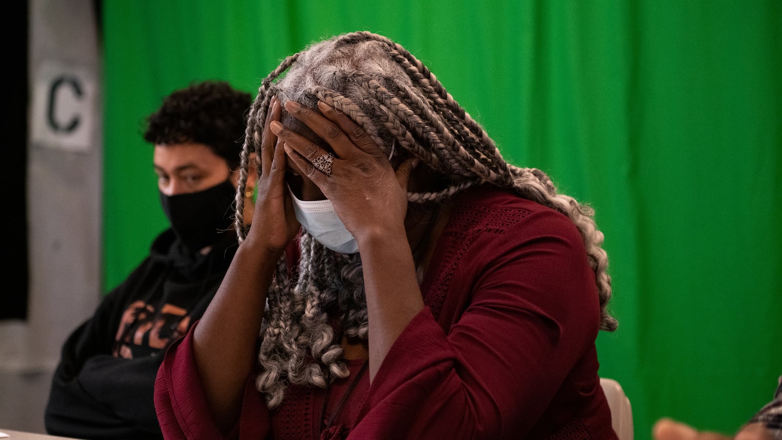A woman with long grey braided hair, wearing a mask and red dress, puts her hands over her head while sitting at a table. Someone sis sitting to the left of the woman and there is a bright green backdrop behind them.
