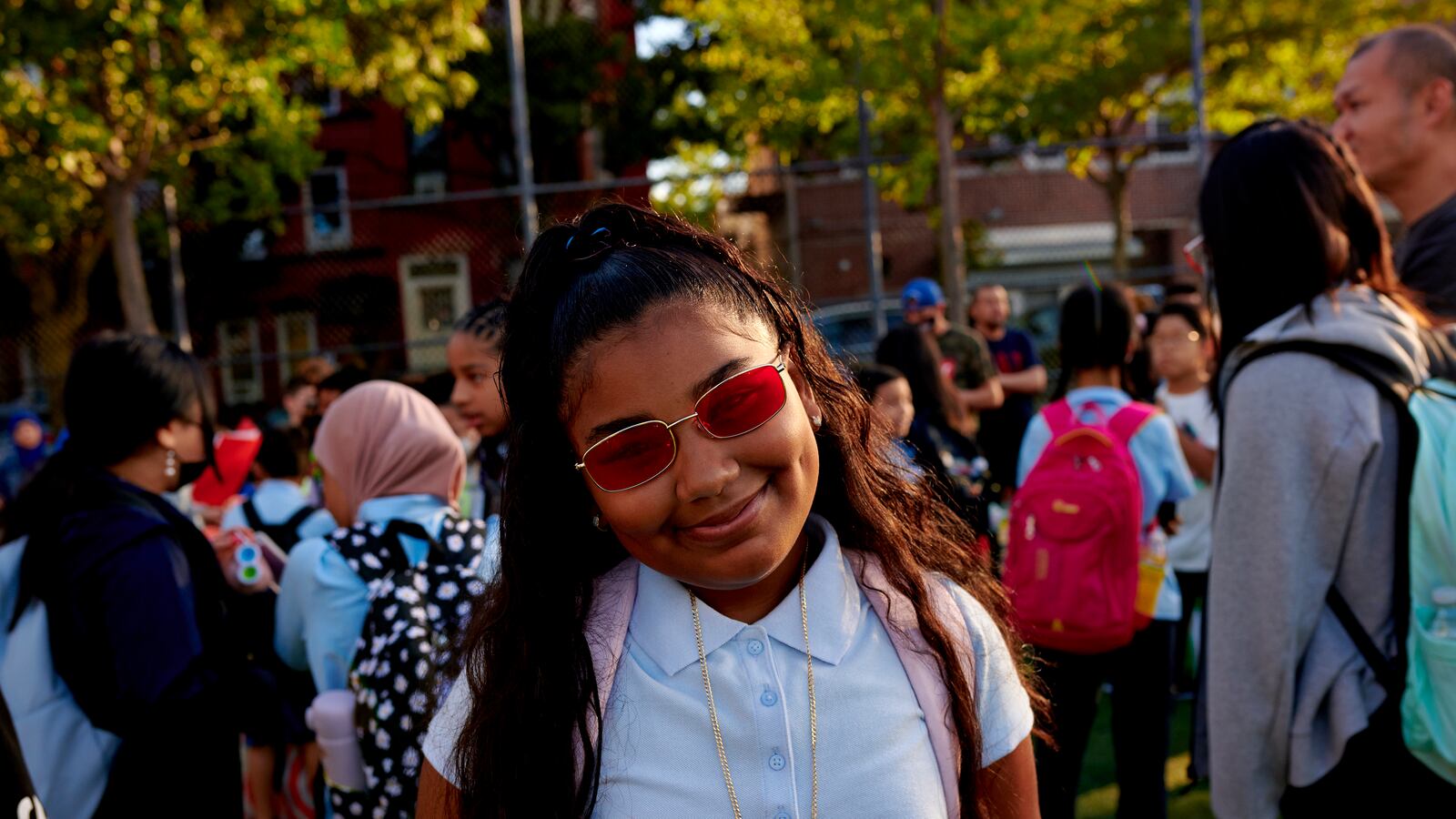 A girl smiles for a portrait, wearing a gold necklace and red sunglasses.