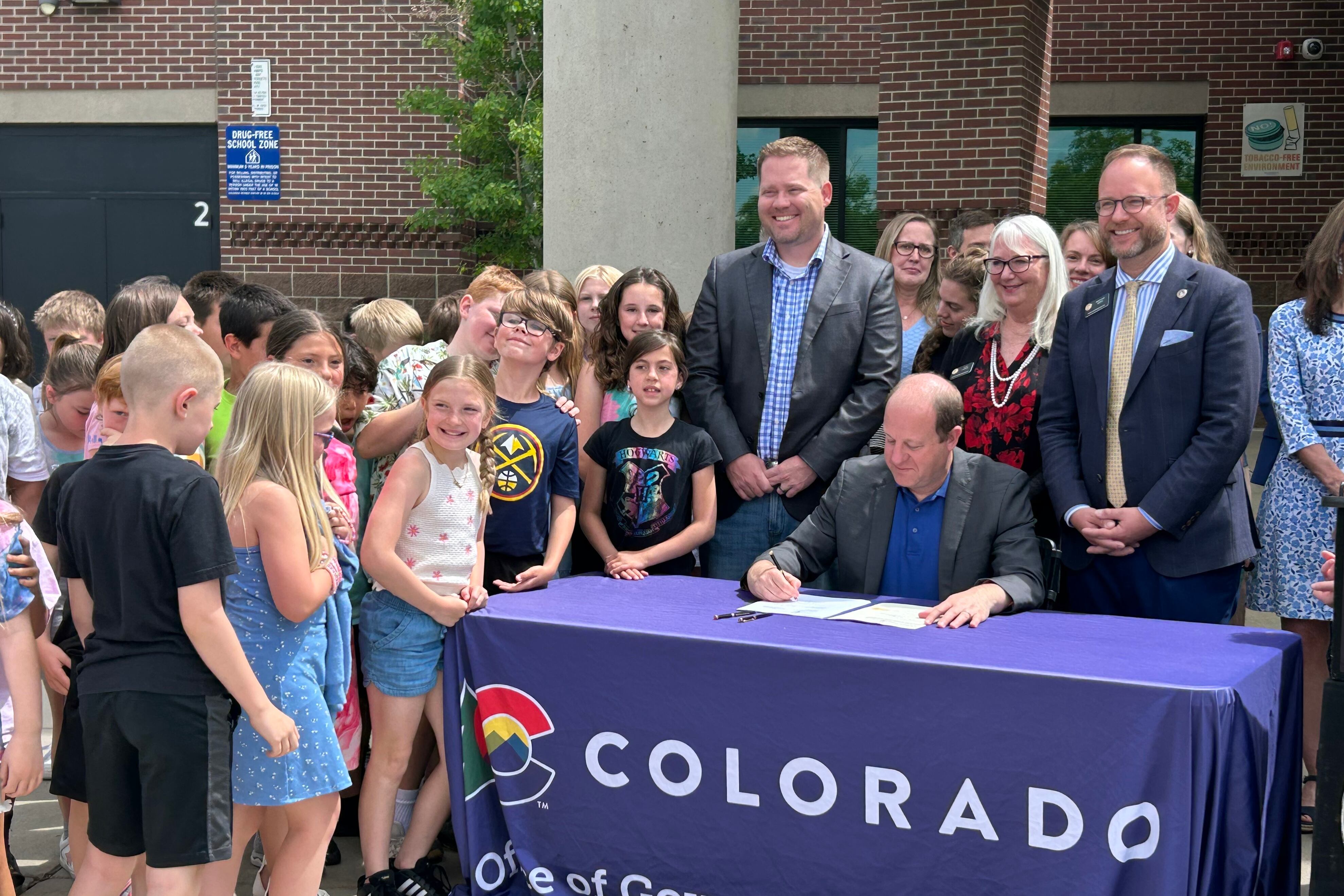 A man in a suit sits at a table with a purple table cloth with adults and children standing around him.