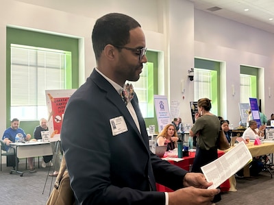 A man wearing a suit holds a pamphlet in a room with tables and people sitting in the background.