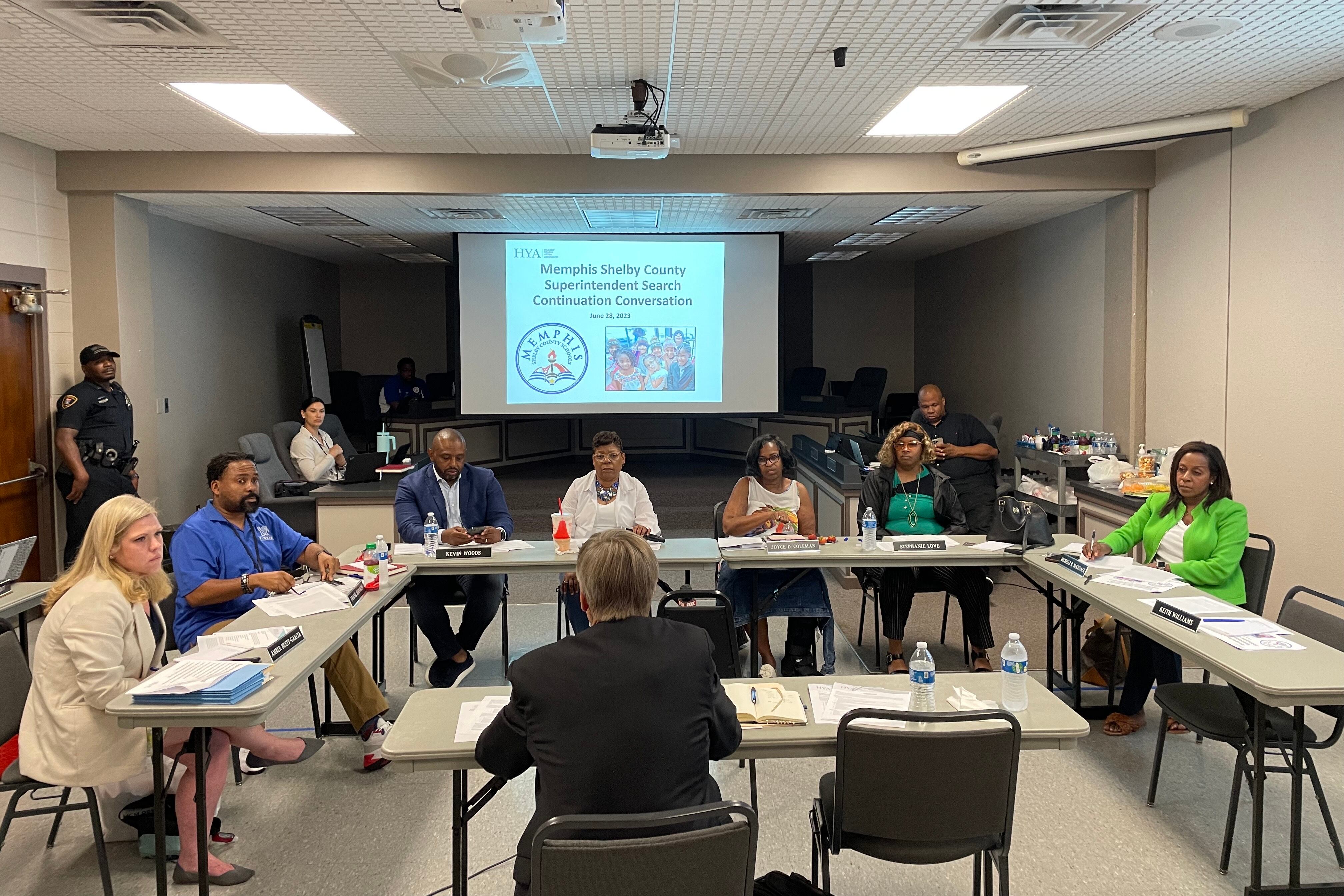 Seven people gather around a set of tables to listen to a man in a suit speak about a presentation called “Memphis Shelby County Superintendent Search Continuation Conversation”