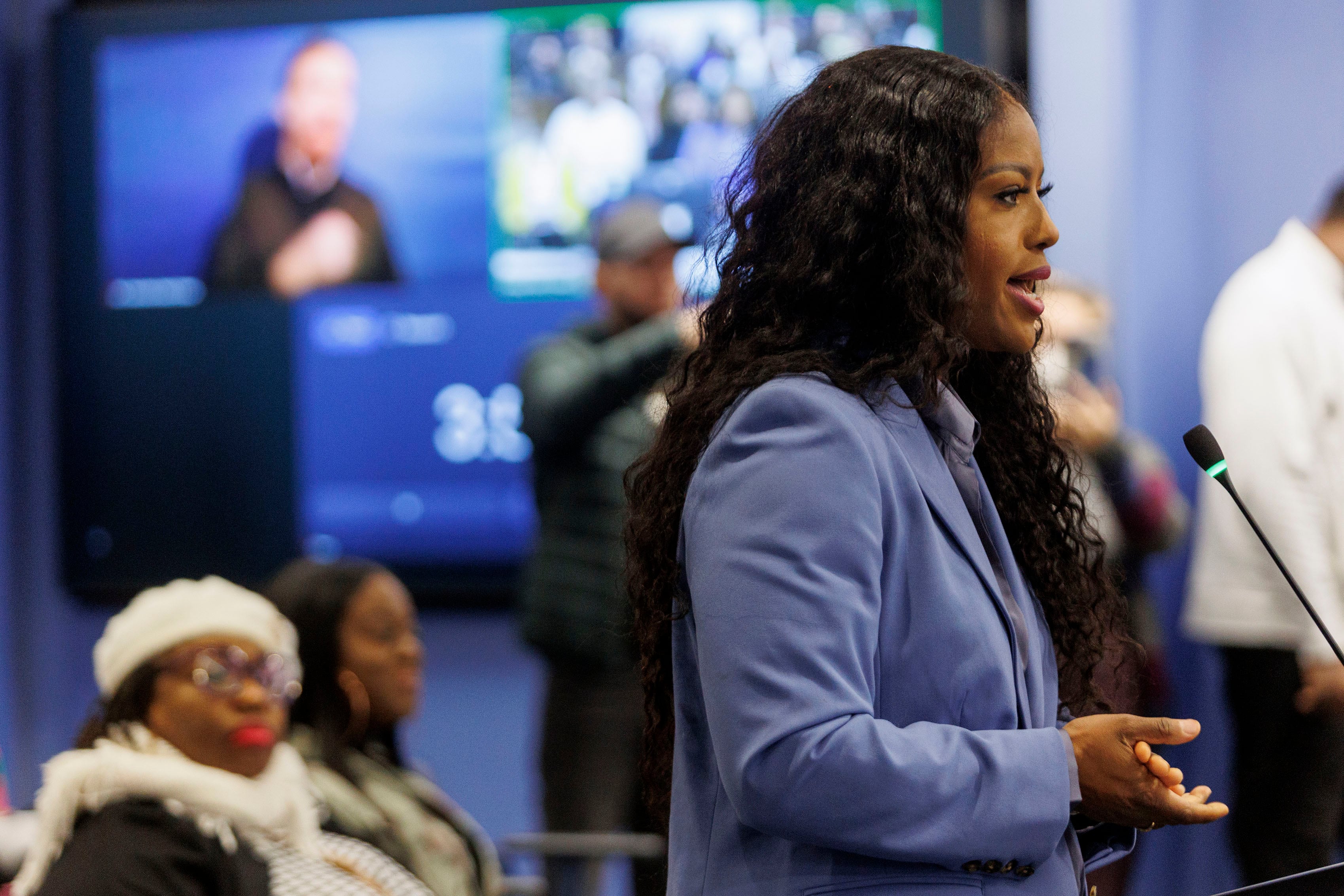 A woman with long, dark and curly hair and wearing a blue suit speaks from behind a podium and in front of a crowd.