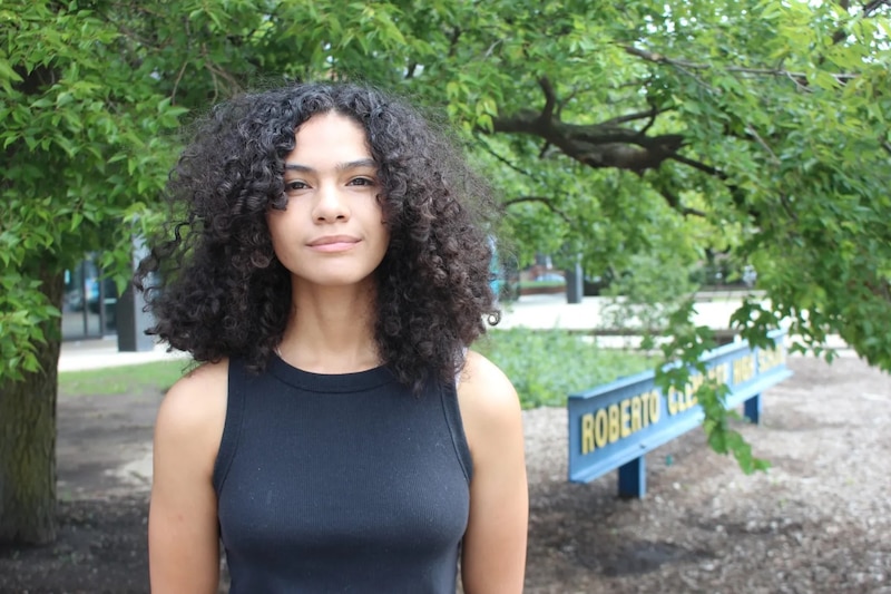 A woman with dark hair and a dark top stands in front of a tree.